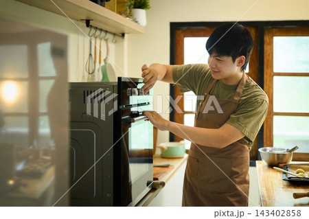Man in Apron Placing Cookie Tray into Oven While Baking Homemade Cookies in Modern Kitchen Man in Apron Placing Cookie Tray into Oven While Baking Homemade Cookies in Modern Kitchen 143402858