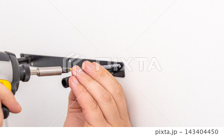 Mans hands using yellow cordless drill with extension bit to secure black bracket on white wall. Home improvement, extended reach fastening, DIY shelf mounting, precise work. Mans hands using yellow cordless drill with extension bit to secure black bracket on white wall. Home improvement, extended reach fastening, DIY shelf mounting, precise work. 143404450