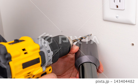 Mans hands holding yellow cordless drill with bit near wall outlet, ready for drilling. Home improvement, preparing to drill, DIY drilling, tool handling. 143404482