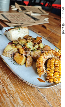 Roasted potato chunks, vegetables, and corn on a plate beside sliced bread and menus on a rustic wooden dining table, close up food scene 143405038
