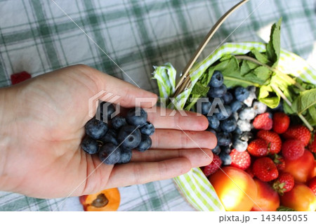 Hand holding fresh blueberries beside a woven basket filled with strawberries, raspberries, apples, tomatoes, and leafy greens on a checked cloth 143405055