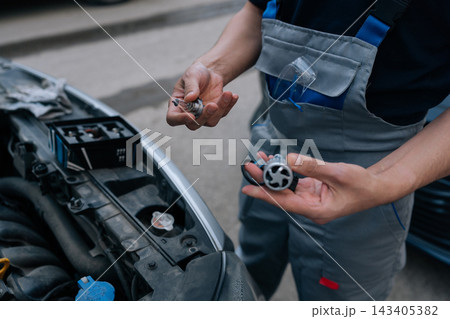Auto mechanic comparing traditional halogen bulb with modern led headlight bulb in hand, standing near car engine bay, preparing for vehicle lighting upgrade or repair. Auto mechanic comparing traditional halogen bulb with modern led headlight bulb in hand, standing near car engine bay, preparing for vehicle lighting upgrade or repair. 143405382