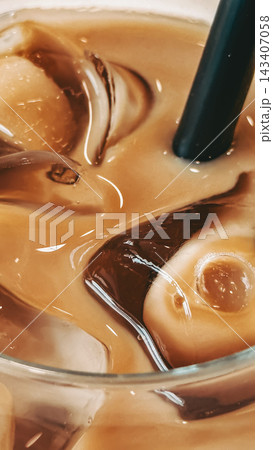Iced coffee with ice cubes and black straw. Iced coffee fills glass with translucent ice cubes and drinking straw. Close view of iced coffee shows creamy brown tones, melting ice, bright reflections Iced coffee with ice cubes and black straw. Iced coffee fills glass with translucent ice cubes and drinking straw. Close view of iced coffee shows creamy brown tones, melting ice, bright reflections 143407058