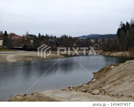 Tranquil overcast shoreline with distant settlement. Silent cloudy water edge surrounded by rocky ground Tranquil overcast shoreline with distant settlement. Silent cloudy water edge surrounded by rocky ground 143407457