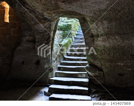 Cave interior stone staircase carved from rock walls moss covered light filtering through opening illuminates rough texture and ancient passageway for adventurous exploration Cave interior stone staircase carved from rock walls moss covered light filtering through opening illuminates rough texture and ancient passageway for adventurous exploration 143407869