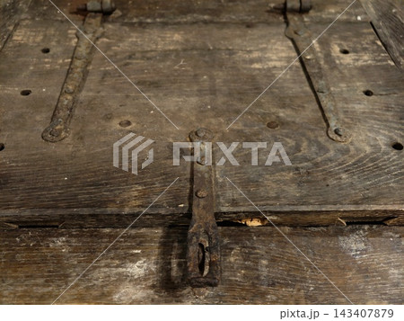 Weathered wooden planks with rusty hinges and aged hardware reveal patina, grain, and worn surface across old boards in dim workshop light, showing cracks, gaps, nails, screws, and metal straps 143407879