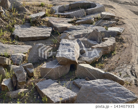 Cracked granite rubble and weathered concrete blocks scattered along sunlit dirt shoulder with tufts grasses and hardy weeds growing in crevices under warm afternoon light Cracked granite rubble and weathered concrete blocks scattered along sunlit dirt shoulder with tufts grasses and hardy weeds growing in crevices under warm afternoon light 143407935