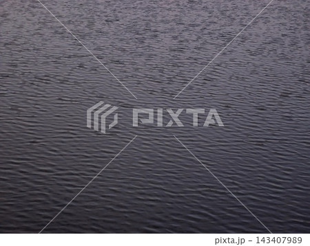 Dusk lit rippled water surface with subtle waves and muted purple blue tones across wide calm expanse conveying tranquil reflective texture and quiet atmosphere Dusk lit rippled water surface with subtle waves and muted purple blue tones across wide calm expanse conveying tranquil reflective texture and quiet atmosphere 143407989