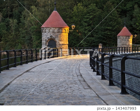 Historic stone towers with red conical roofs overlook cobblestone riverside walkway lined with lampposts glowing at dusk, stone bridge visible beyond river nearby 143407993