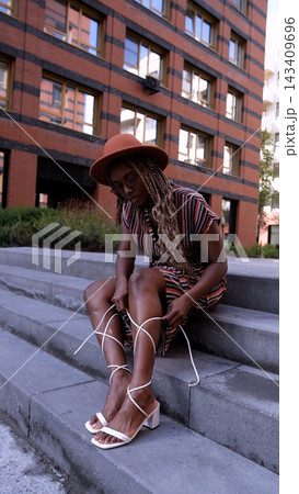 A beautiful young woman with afro braids and a brown hat poses while sitting on the steps outside in an eco-friendly residential complex. She ties the laces of her sandals and smiles, looking at the A beautiful young woman with afro braids and a brown hat poses while sitting on the steps outside in an eco-friendly residential complex. She ties the laces of her sandals and smiles, looking at the 143409696