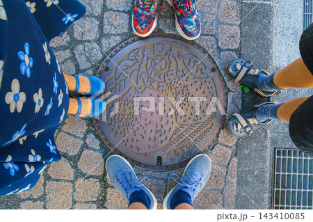Yatsushiro, Kumamoto Pref, Japan - Oct 9 2024, close-up view of a manhole cover with a picture of a melon on it, surrounded by the feet of a family of four, at daytime, Yatsushiro, Japan Yatsushiro, Kumamoto Pref, Japan - Oct 9 2024, close-up view of a manhole cover with a picture of a melon on it, surrounded by the feet of a family of four, at daytime, Yatsushiro, Japan 143410085