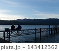 湖の桟橋に座り山並みを眺める二人 143412362