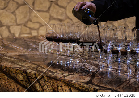 Hand pouring red wine from a bottle into tasting glasses on a beautiful marble counter, preparing for an exclusive winery event or vintage barrel aging sampling 143412929