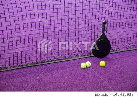 Black padel racket leaning against mesh net on purple turf with three fluorescent balls, copy space Black padel racket leaning against mesh net on purple turf with three fluorescent balls, copy space 143413938