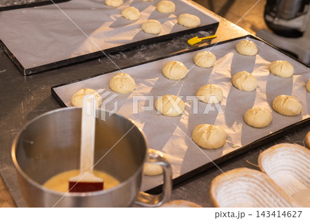 Two parchment-lined trays holding scored rolls are proofing on stainless counter with pastry brush Two parchment-lined trays holding scored rolls are proofing on stainless counter with pastry brush 143414627