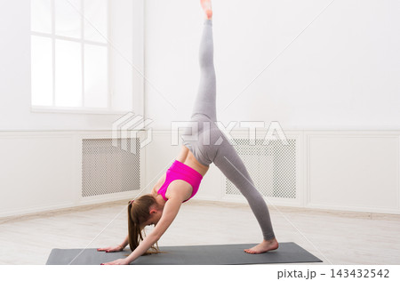 A woman is doing a downward dog pose on a mat in a bright studio. She is focused on her balance and stretching. Natural light fills the space through large windows. 143432542