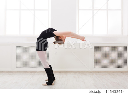 A dancer performs a backbend in a bright studio. The dancer wears black and white clothing. Natural light enters through the large windows, illuminating the space. 143432614