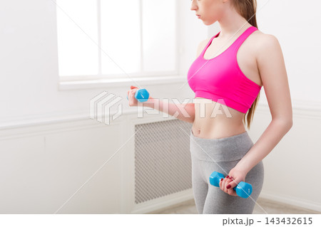 A woman stands in a fitness studio holding blue dumbbells. She focuses on her workout while wearing a pink top and gray pants. Light comes in from a window nearby. 143432615