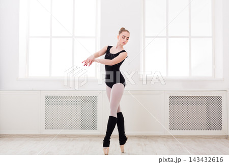 A ballet dancer is performing in a bright studio with large windows letting in natural light. She wears a black outfit and shows her skills with an elegant pose, focused on her movements. A ballet dancer is performing in a bright studio with large windows letting in natural light. She wears a black outfit and shows her skills with an elegant pose, focused on her movements. 143432616