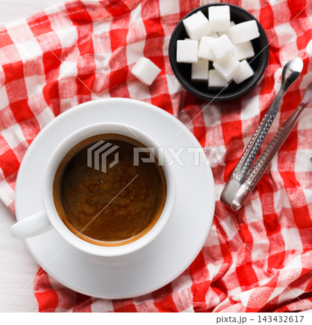 A cup of coffee sits on a white saucer next to a small bowl of sugar cubes. The red checkered tablecloth adds a touch of color to the scene. Tongs rest beside the bowl for serving the sugar. A cup of coffee sits on a white saucer next to a small bowl of sugar cubes. The red checkered tablecloth adds a touch of color to the scene. Tongs rest beside the bowl for serving the sugar. 143432617