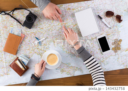 Two people are gathered around a map on a wooden table. They are discussing travel plans while enjoying coffee. Nearby, there are travel documents, a camera, and a smartphone. Two people are gathered around a map on a wooden table. They are discussing travel plans while enjoying coffee. Nearby, there are travel documents, a camera, and a smartphone. 143432758