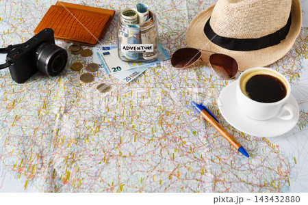 Items are laid out on a map for travel planning. A camera, money, coins, a notebook, sunglasses, a hat, and coffee are present. Elements suggest preparation for an adventure or trip. Items are laid out on a map for travel planning. A camera, money, coins, a notebook, sunglasses, a hat, and coffee are present. Elements suggest preparation for an adventure or trip. 143432880