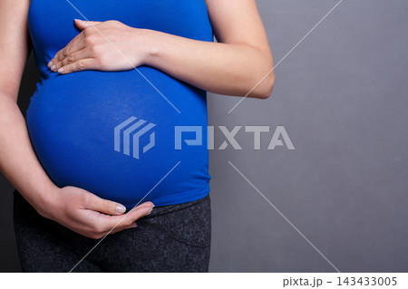 A person is standing against a plain background while holding their pregnant belly. They are wearing a blue top and gray pants, and appear to be in a moment of reflection. A person is standing against a plain background while holding their pregnant belly. They are wearing a blue top and gray pants, and appear to be in a moment of reflection. 143433005