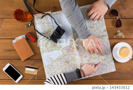 Two people examine a map on a wooden table while planning their trip. Nearby are a camera, coins, a phone, and a cup of coffee. Items show preparation for a journey together and exploration. 143433012