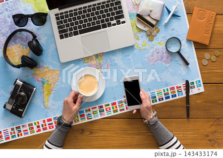 A person sits at a wooden table with a laptop, map, coffee cup, smartphone, and travel items. Various currencies and a camera are scattered around, indicating trip preparation. 143433174
