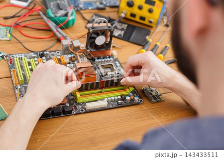 A person is focused on assembling parts of a computer motherboard in a workshop. Tools and components are scattered on the table around them. The setting is busy with various electronic devices. 143433511