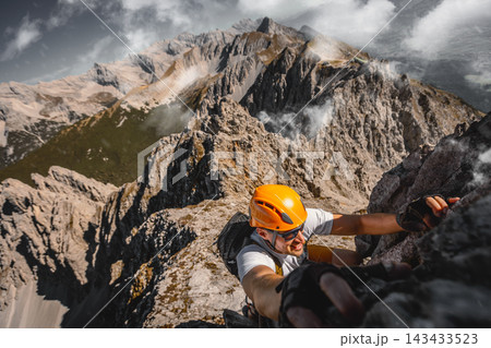 Close-up of a focused solo male climber ascending a rugged rock wall on a via ferrata in the Austrian Alps near Innsbruck, using safety equipment and climbing gear. Close-up of a focused solo male climber ascending a rugged rock wall on a via ferrata in the Austrian Alps near Innsbruck, using safety equipment and climbing gear. 143433523