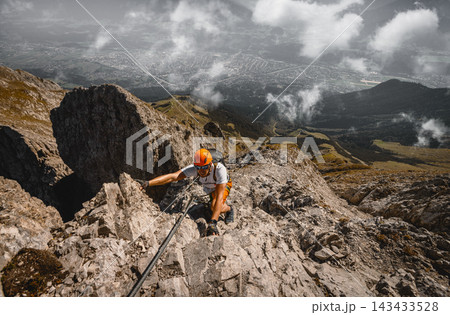 Solo male climber ascending a rocky via ferrata high above an alpine valley near Innsbruck, Austria, surrounded by dramatic mountain scenery, clouds and distant landscapes. 143433528