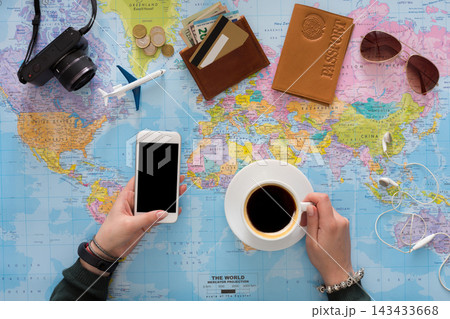 A person holds a smartphone and a cup of coffee while sitting at a table with a world map. Travel items like a passport, wallet, and camera are also present on the table. A person holds a smartphone and a cup of coffee while sitting at a table with a world map. Travel items like a passport, wallet, and camera are also present on the table. 143433668