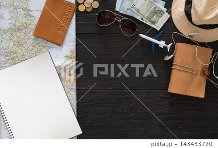 A map lays on a wooden table with a passport, money, sunglasses, a small airplane, and a notebook. Travel items are arranged for planning a trip. Accessories are present for convenience. A map lays on a wooden table with a passport, money, sunglasses, a small airplane, and a notebook. Travel items are arranged for planning a trip. Accessories are present for convenience. 143433720