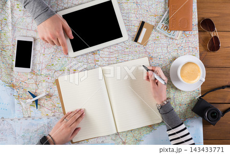 Two people are gathered at a table with a map spread out. They are using devices and writing notes. A cup of coffee and other travel items are on the table. Two people are gathered at a table with a map spread out. They are using devices and writing notes. A cup of coffee and other travel items are on the table. 143433771