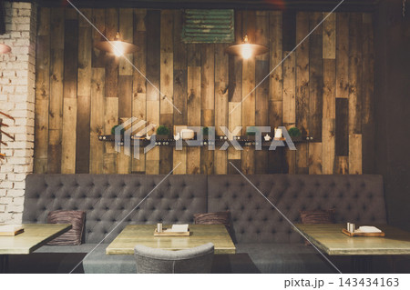 A collection of wooden planks forms a wall in a restaurant. The seating area shows dark gray sofas and wooden tables. Simple light fixtures hang from the ceiling. A shelf holds plants and candles. A collection of wooden planks forms a wall in a restaurant. The seating area shows dark gray sofas and wooden tables. Simple light fixtures hang from the ceiling. A shelf holds plants and candles. 143434163