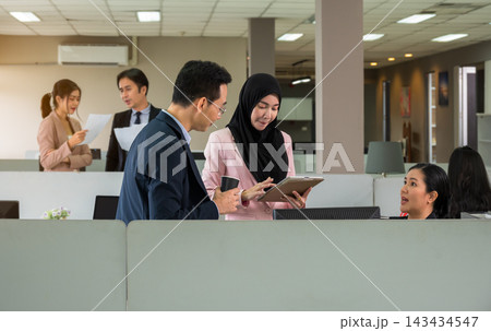 Diversity group of colleagues in open space office. Woman in hijab sharing information on digital tablet with Asian businessman while others working and interacting in background Diversity group of colleagues in open space office. Woman in hijab sharing information on digital tablet with Asian businessman while others working and interacting in background 143434547