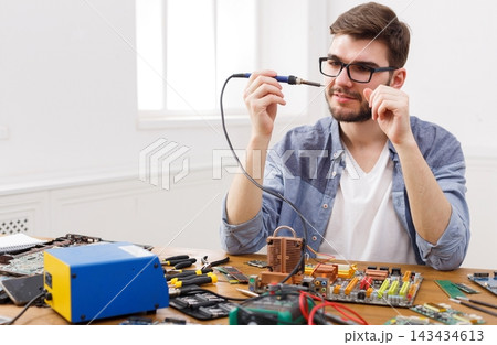 A man is sitting at a table focused on fixing electronic devices. He is using a soldering iron while surrounded by various tools and parts. The room has plenty of light. A man is sitting at a table focused on fixing electronic devices. He is using a soldering iron while surrounded by various tools and parts. The room has plenty of light. 143434613