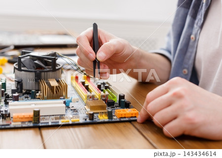 A person is focused on assembling or repairing a computer motherboard using tweezers. Tools are spread on a wooden table in a bright workshop setting. A person is focused on assembling or repairing a computer motherboard using tweezers. Tools are spread on a wooden table in a bright workshop setting. 143434614