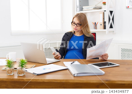 A pregnant woman sits at a wooden desk in a bright office. She holds a phone in one hand and papers in the other, focused on her work. The laptop is open and small plants are on the desk. 143434977