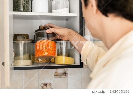 man reaches for a glass jar of red lentils in the organized cabinet. Shelves hold cereals, rice, couscous, and healthy grains stored cleanly in neat glass containers for cooking. 143435193