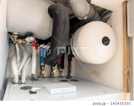 A new white expansion vessel with pipes and brass fittings under an open boiler. Servicing a hot water installation A new white expansion vessel with pipes and brass fittings under an open boiler. Servicing a hot water installation 143435335