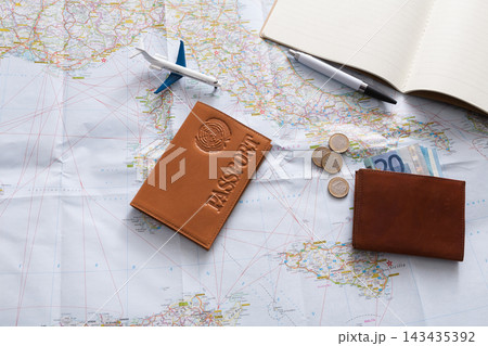 A workspace shows a passport, coins, and cash placed on a map. An airplane model and a pen are also on the table. This scene suggests travel planning and preparation. A workspace shows a passport, coins, and cash placed on a map. An airplane model and a pen are also on the table. This scene suggests travel planning and preparation. 143435392