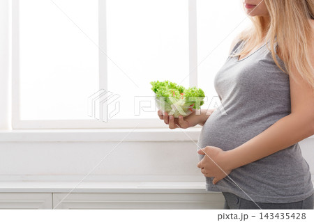 A pregnant woman stands by a window holding a bowl of fresh lettuce. She is in a light room with white walls. 143435428