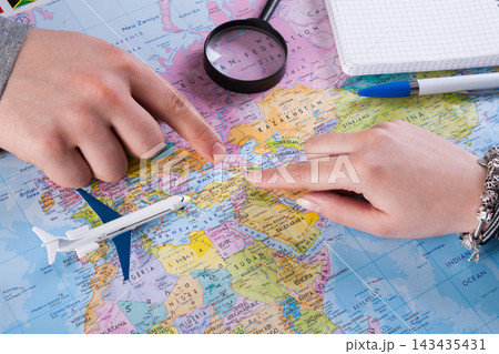 Two hands point at different locations on a map while planning a trip. A magnifying glass is nearby, along with a notepad and pens. An airplane model is placed on the map. Two hands point at different locations on a map while planning a trip. A magnifying glass is nearby, along with a notepad and pens. An airplane model is placed on the map. 143435431