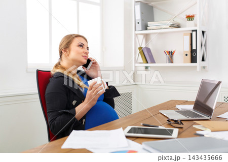 A pregnant woman is sitting at a desk in her home office. She is on the phone and holding a cup. Papers and a laptop are on the desk. The room has natural light from a window. A pregnant woman is sitting at a desk in her home office. She is on the phone and holding a cup. Papers and a laptop are on the desk. The room has natural light from a window. 143435666