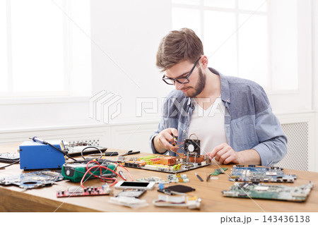 A person is focused on repairing a circuit board on a wooden table. Various electronic components and tools are spread around. The room has large windows that let in natural light. A person is focused on repairing a circuit board on a wooden table. Various electronic components and tools are spread around. The room has large windows that let in natural light. 143436138
