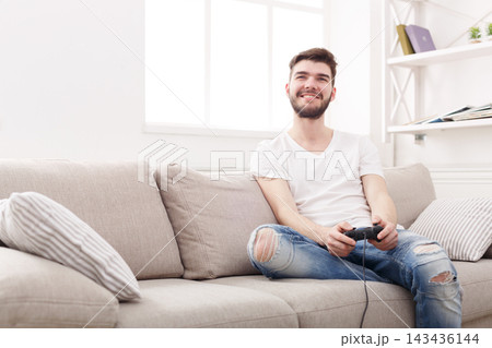 A man is enjoying a video game on a couch. He holds a game controller in his hands and smiles. The living room has bright light coming from a window nearby. 143436144