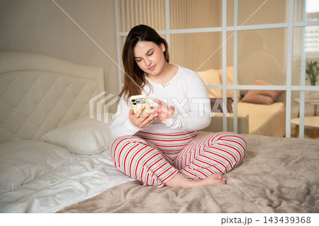 Young woman checking smartphone notifications while sitting on bed in a bright bedroom at home. Morning mobile routine digital communication and leisure lifestyle Young woman checking smartphone notifications while sitting on bed in a bright bedroom at home. Morning mobile routine digital communication and leisure lifestyle 143439368