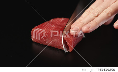 Hand slicing fresh raw tuna steak with a sharp knife on a dark background for gourmet seafood preparation 143439850